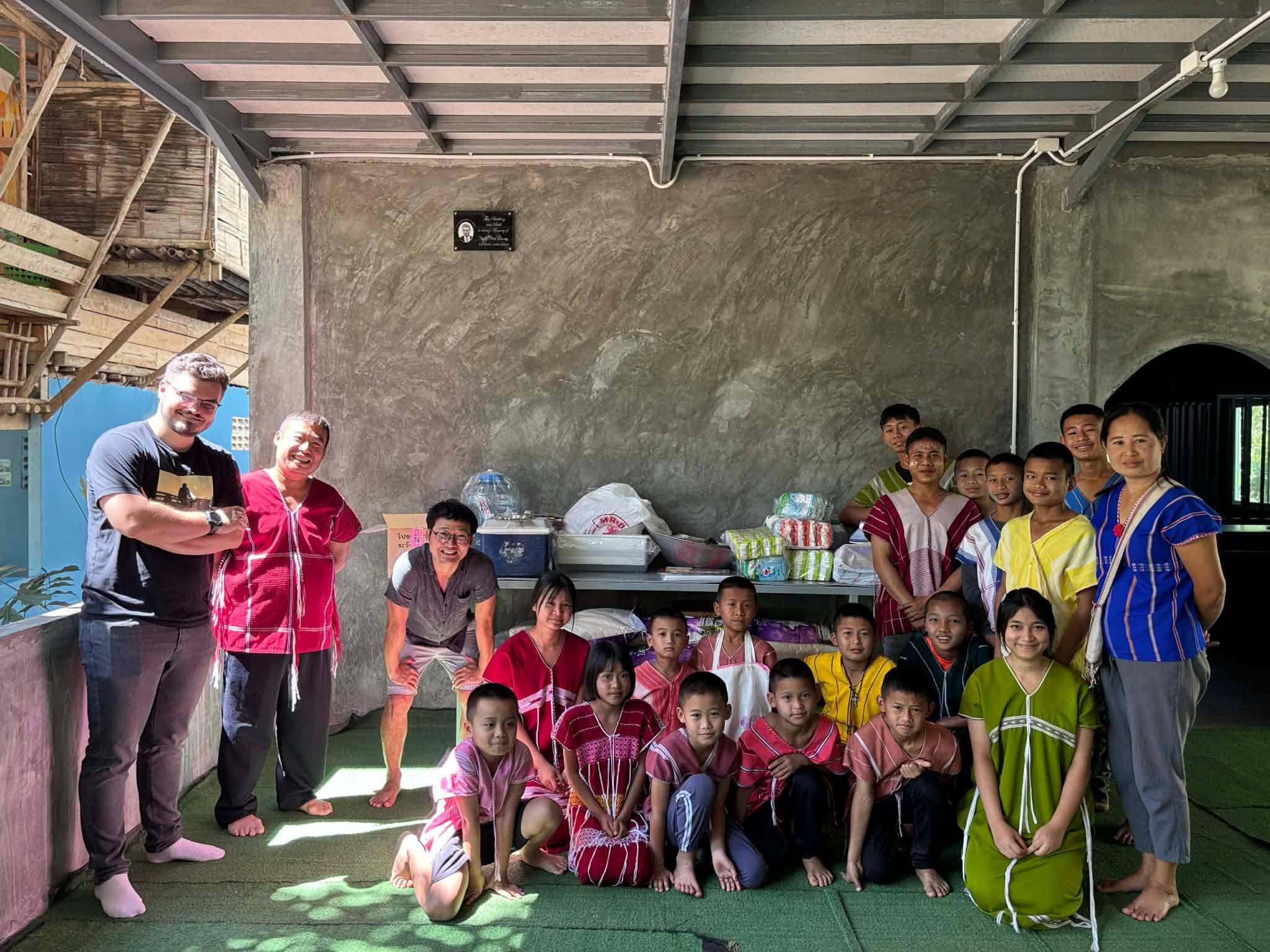 Rice Run 4 · February 2026 — Children in traditional Karen dress and adults posed in a concrete room with supplies stacked on a table behind them
