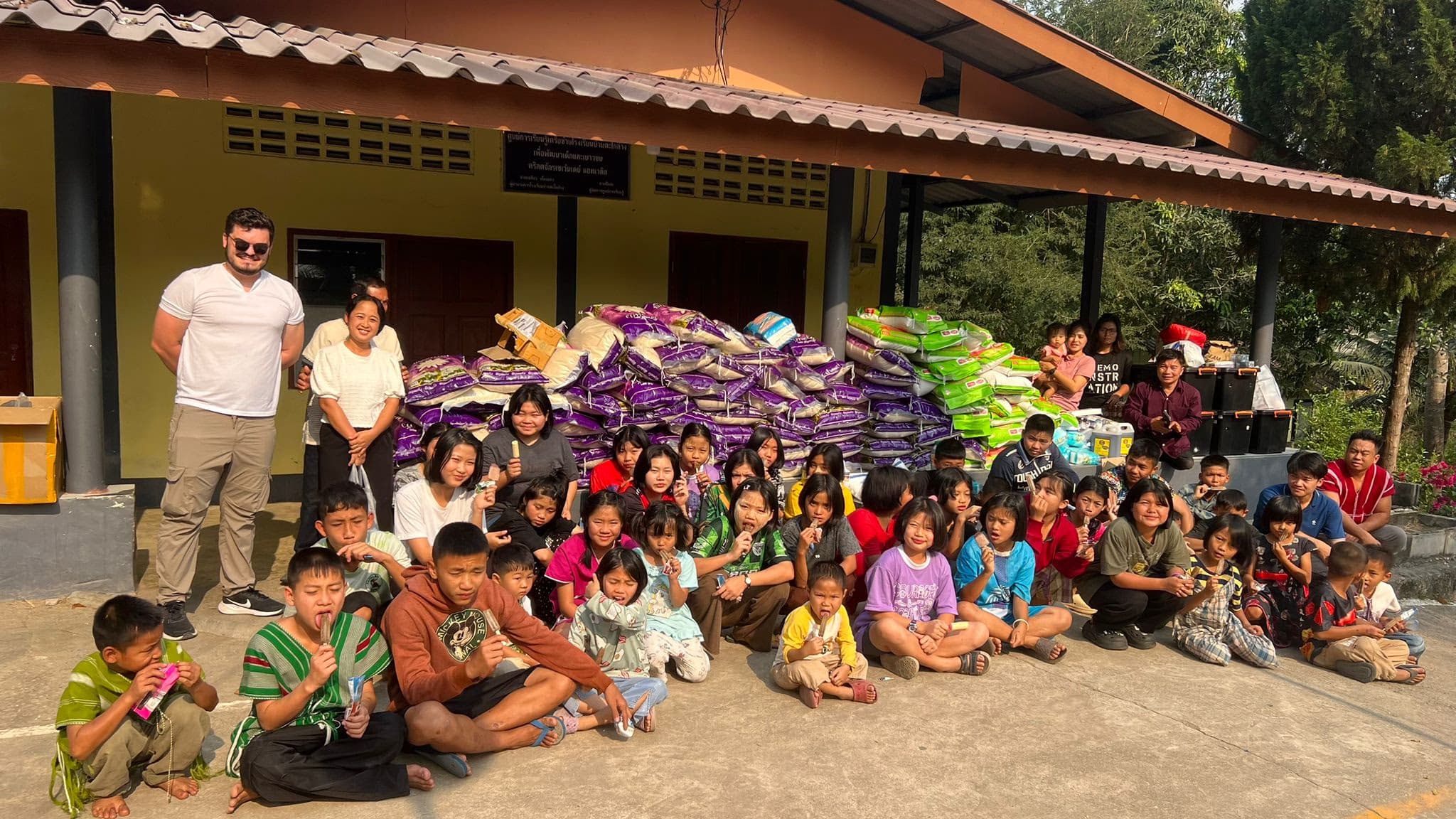 Rice Run 4 · February 2026 — Around forty children and adults sitting and standing in front of a yellow building with a large pile of rice sacks behind them