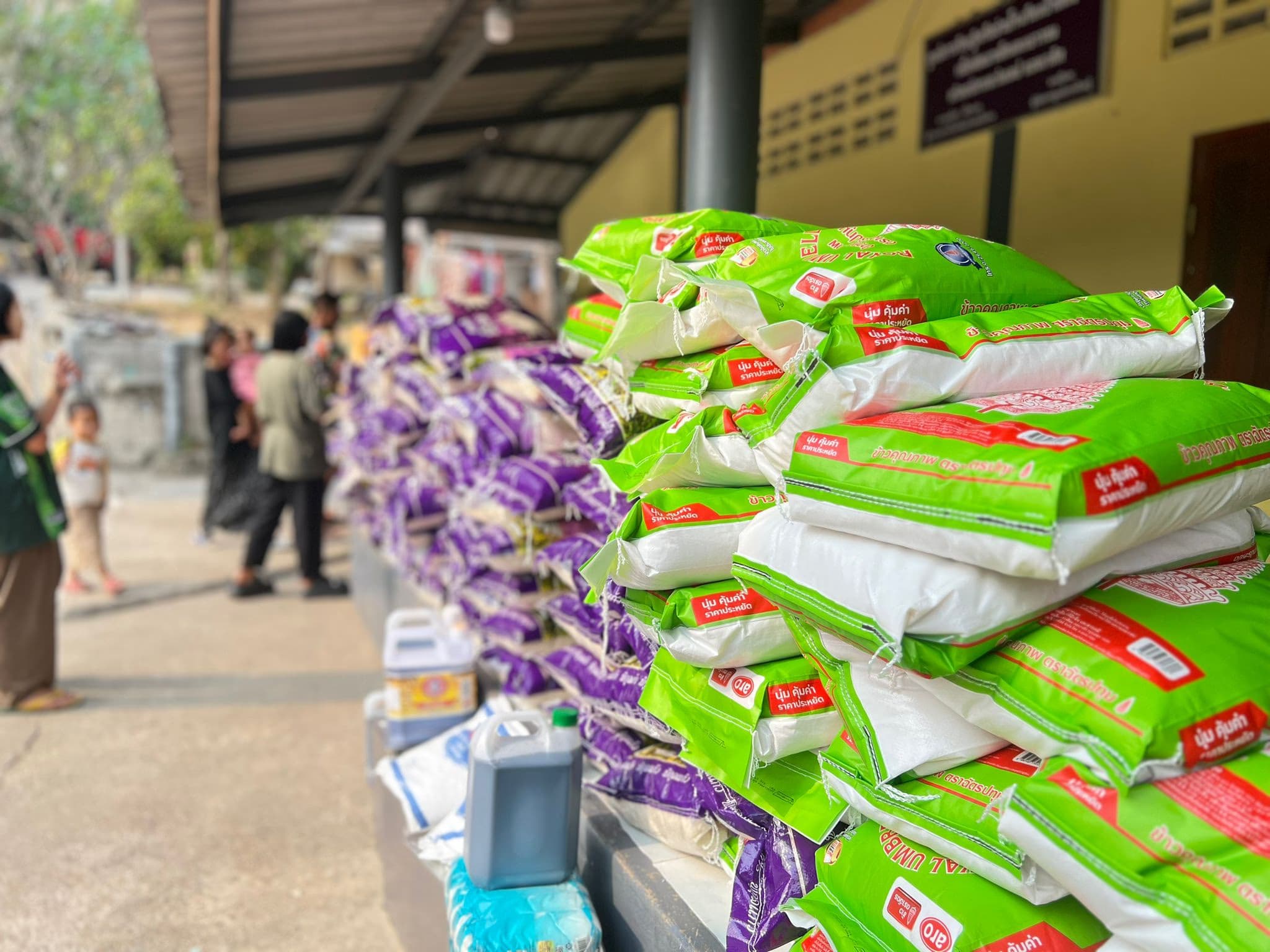 Close view of stacked green and purple sacks of rice with jerrycans of cooking oil at the base, people out of focus behind