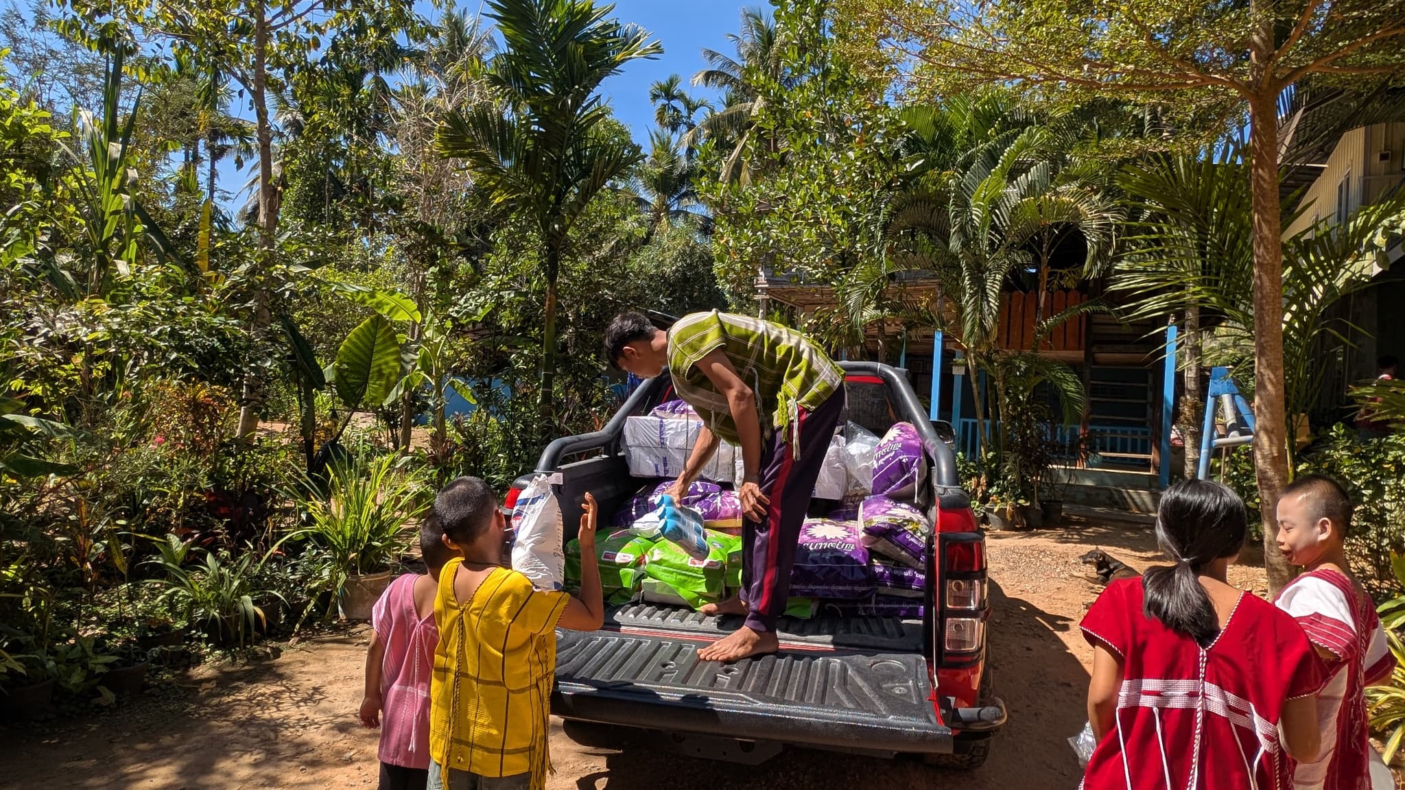 A man standing in a truck bed passing supplies down to children in a jungle yard