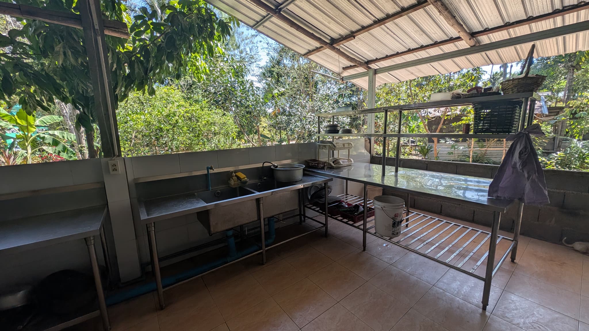 An open-sided kitchen with steel prep tables, a sink, and blackened pots on a shelf, jungle visible through the gap