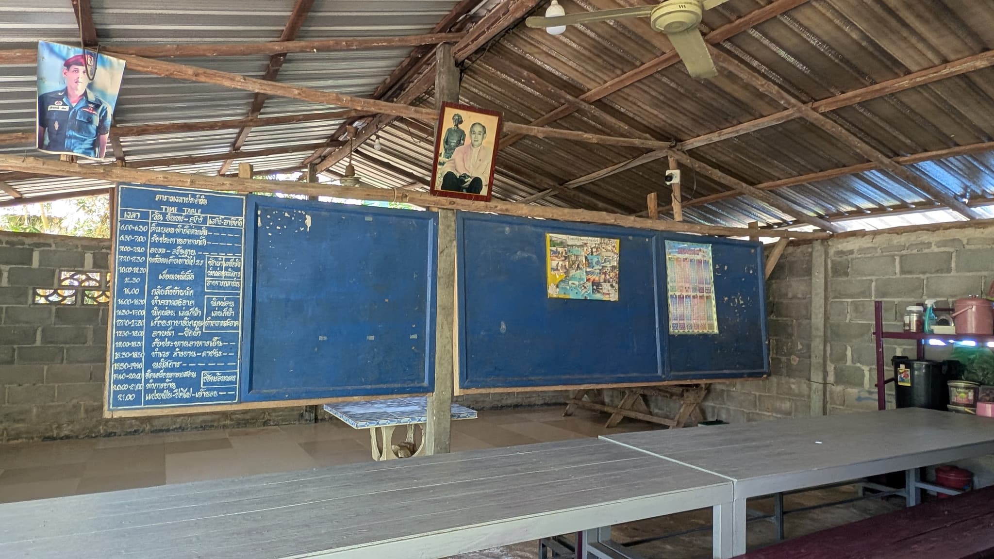A classroom under a corrugated tin roof with three blue blackboards, a handwritten timetable and long wooden benches