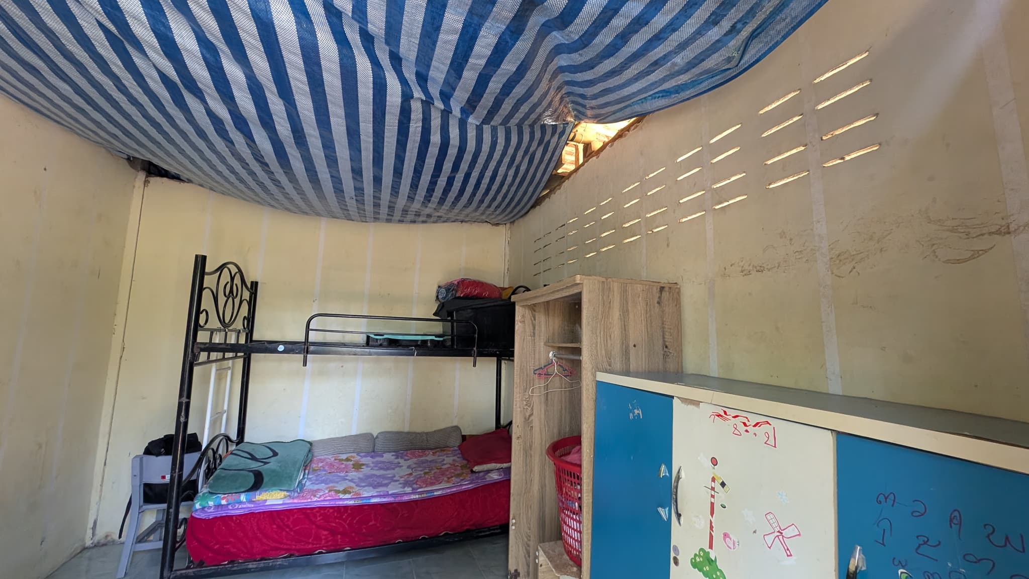 A dormitory room with a metal bunk bed, a wooden cupboard and a striped tarpaulin stretched across the ceiling
