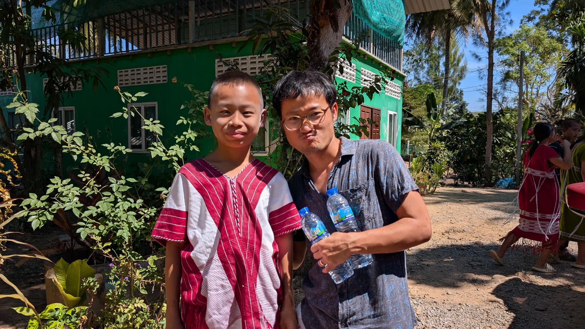 A boy in a red and white traditional Karen shirt standing beside a man, a green building and trees behind them