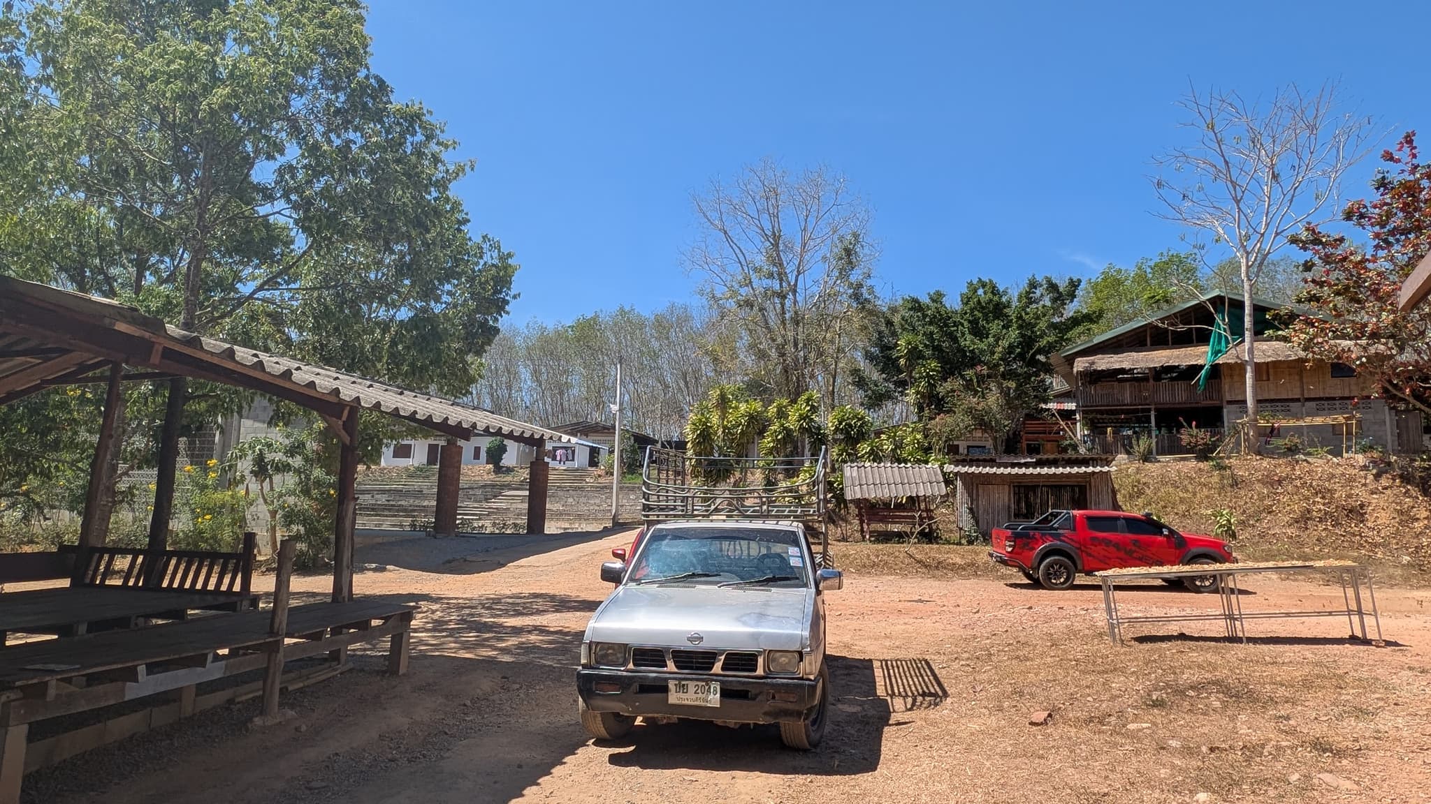 A red dirt clearing with timber buildings under corrugated steel roofs, two parked pickups and dry-season trees under a hard blue sky