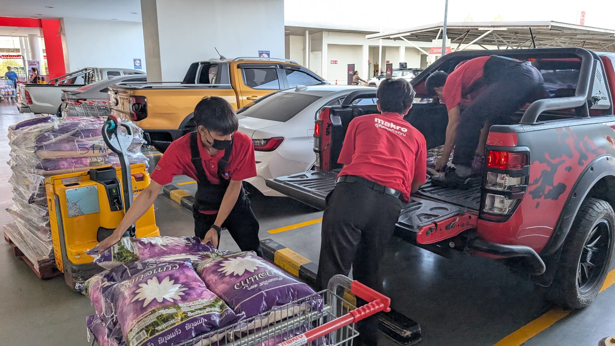 Two staff in red uniforms lifting sacks of rice from a pallet truck into the back of a pickup in a car park