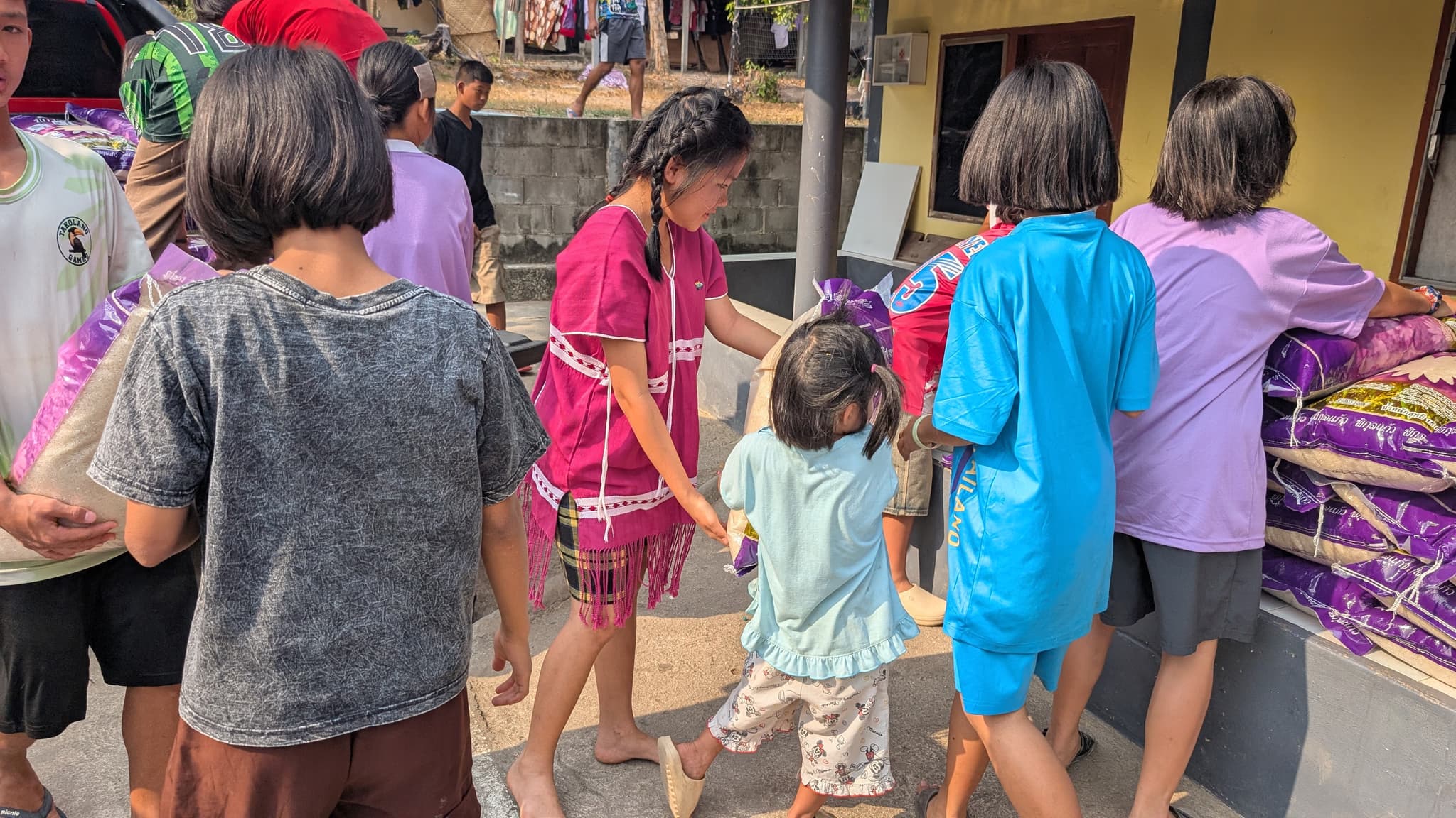 Children walking away from a truck with sacks of rice on their shoulders, a woman in a pink Karen dress leading a small child by the hand