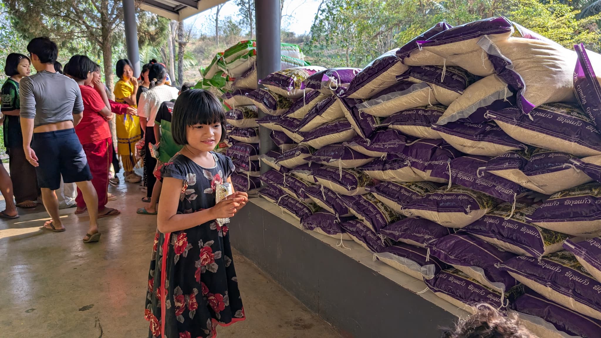 A girl in a floral dress standing beside a wall of stacked purple and white rice sacks under a covered walkway