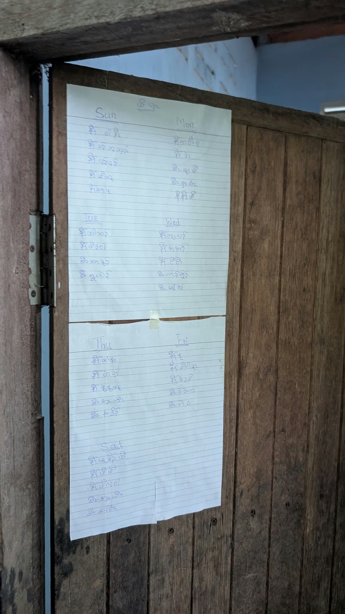 A handwritten meal rota on lined paper taped to a weathered wooden pantry door