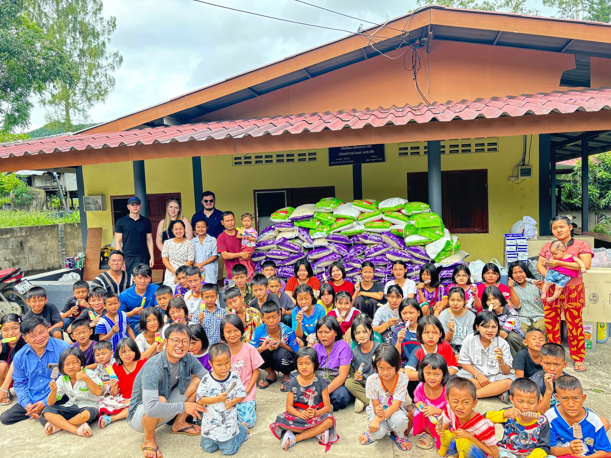 Rice Run 3 · June 2025 — A large group of children seated on the ground in front of a tile-roofed building with a pile of purple and green rice sacks behind them