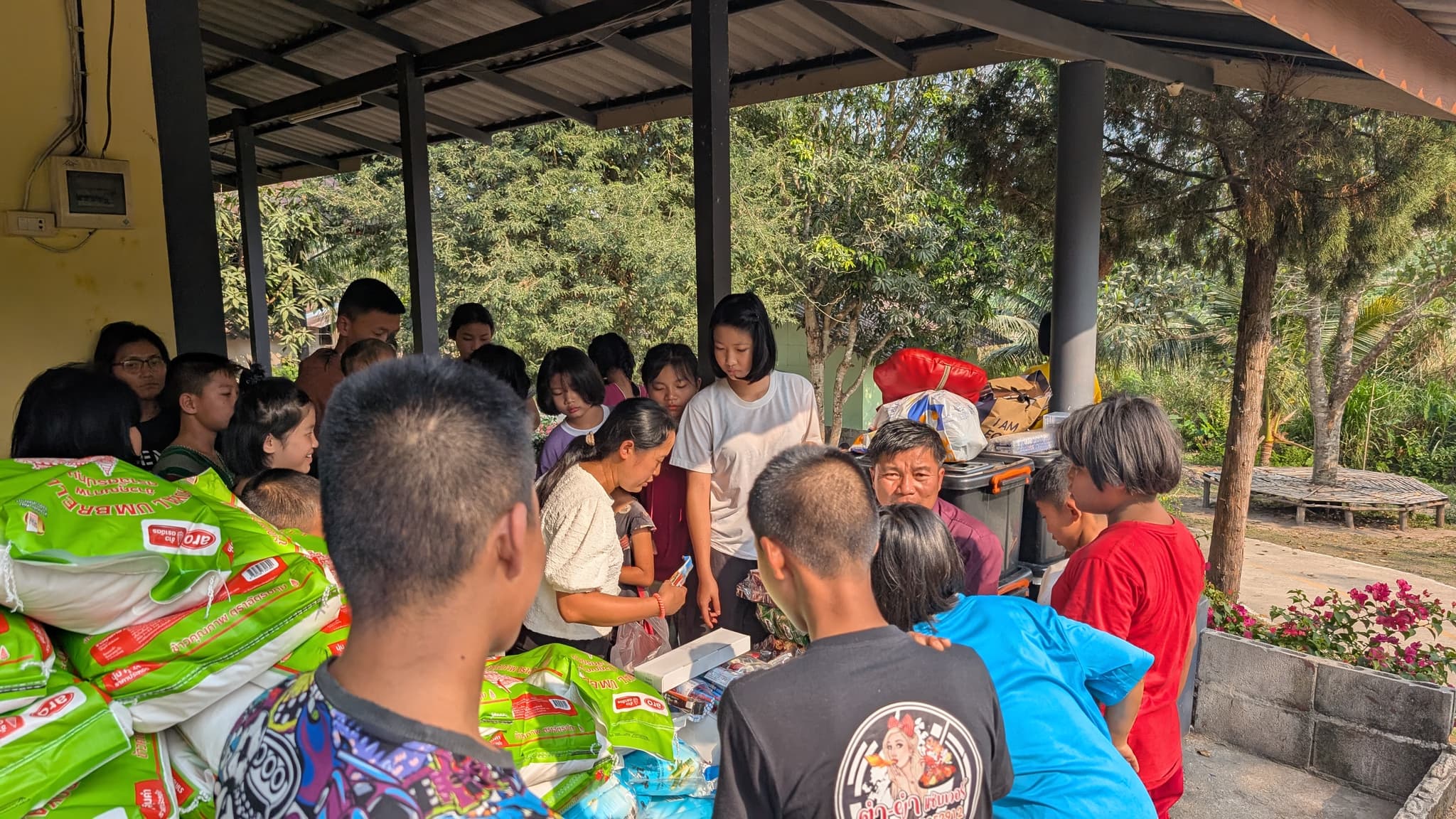 A group of children and adults crowded around a table under a tin-roofed shelter while supplies are handed out, sacks of rice stacked to one side